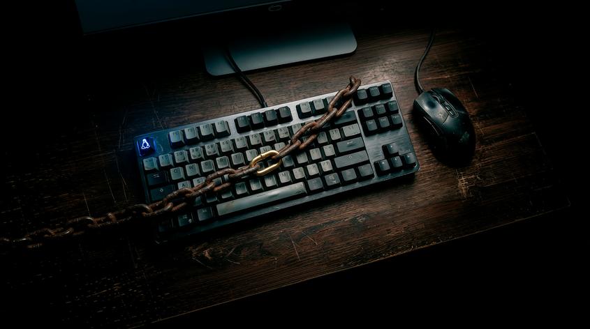A top-down, moody shot of a worn mechanical keyboard on a dark wooden desk. A thick, rusted iron chain with one polished golden link is draped across the keys. The escape key glows with a blue Linux Tux penguin logo. A plain black, well-used mouse sits to the right, and a computer monitor is partially visible in the dark background.
